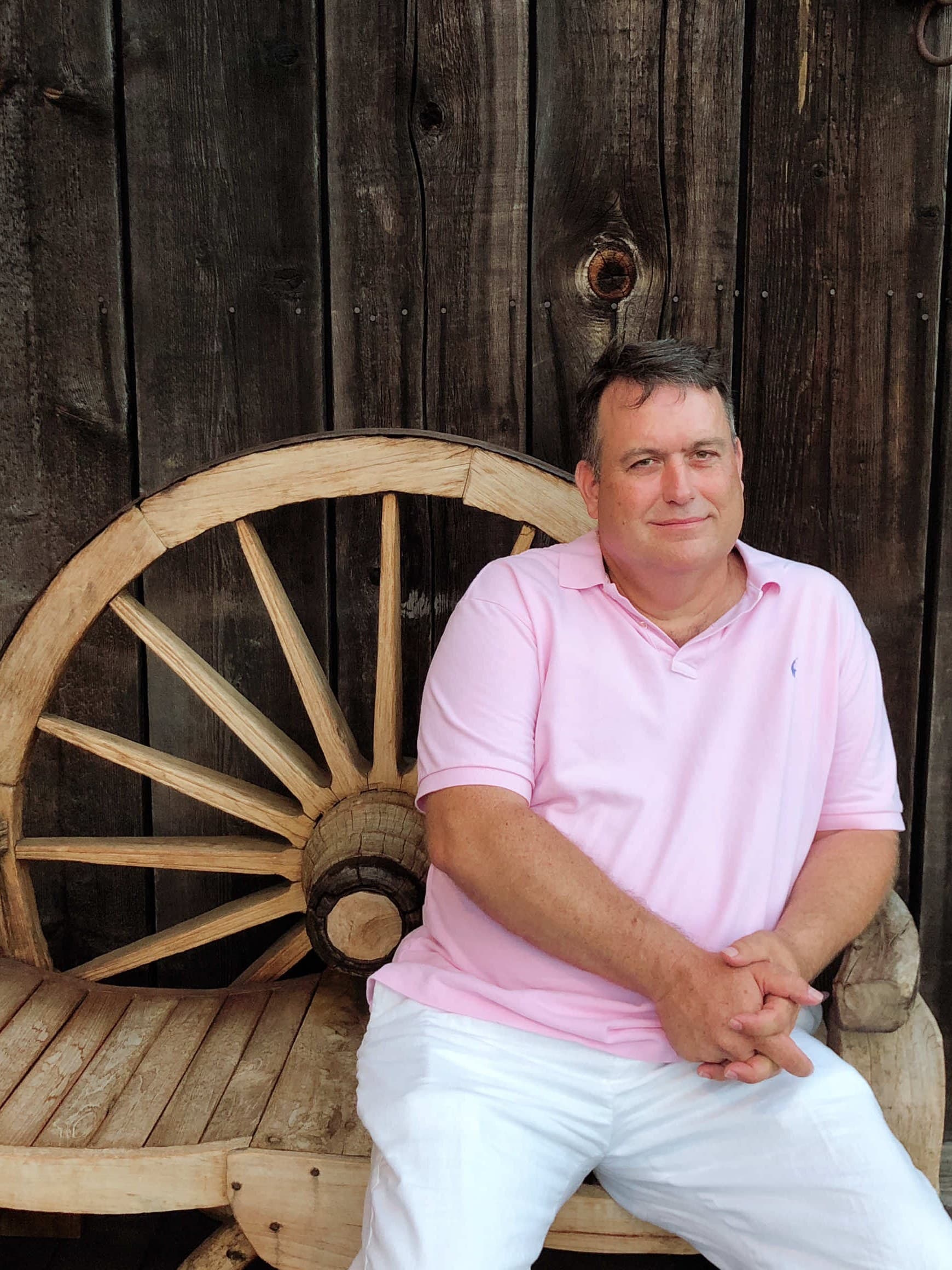 Founder of Florida Pets Aftercare, sitting on a wooden bench with a wagon wheel against a weathered Florida barn wall.